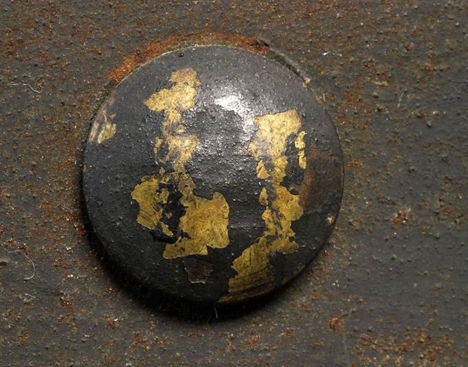 The head of a brass rivet. Note the shape of the head with a more domed (higher) profile. The zinc coating has partly chipped off taking with it the paint. During wear, the rivet heads often lost the coating and the overlaying paint didn’t adhere well to the rivet head’s surface.