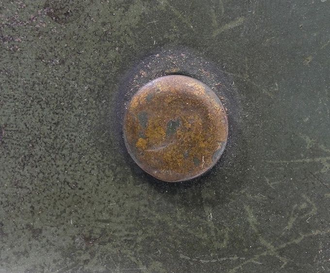 The flat head of a brass rivet. The coating and paint have chipped off. 