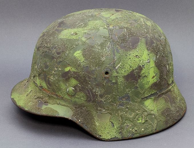 M40 ET64 with sprayed light and dark green in patterns over the surface. Before spraying the helmet received a form of running plasterwork, a unique camouflage scheme. Notice the traces of a former wire attachment. There are at least 4 other known examples in collections in Norway with the same plasterwork pattern, but with different colors.